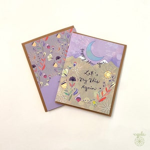 greeting cards with a mountain theme. The front of the card on top has a crescent moon and the words 'Happy New Year Let's Try This Again.' The front of the card on the bottom has a floral pattern with a purple background. funny new year greeting cards