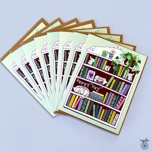 May include: A stack of greeting cards with a whimsical bookshelf design. Each card features a detailed illustration of a bookshelf filled with books, plants, and a sleeping cat. The words "Happy Day" are written on the card.