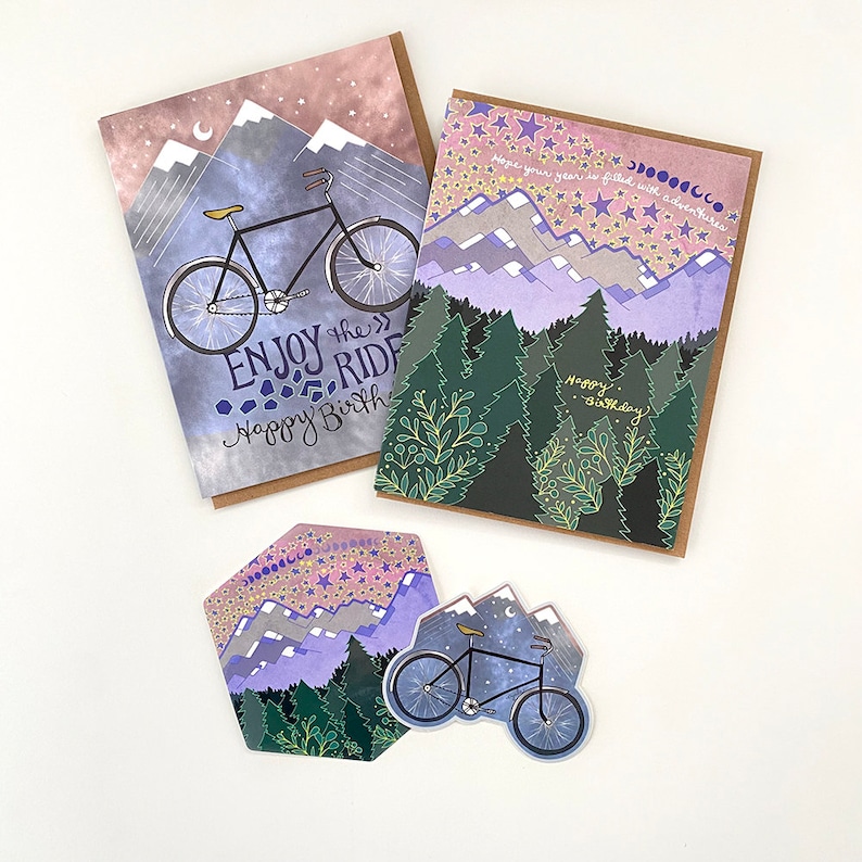 May include: Set of four greeting cards featuring a bicycle, mountains, and stars. The cards are printed on a textured paper with a watercolor effect. The text on the cards reads "Enjoy the Ride" and "Happy Birthday".