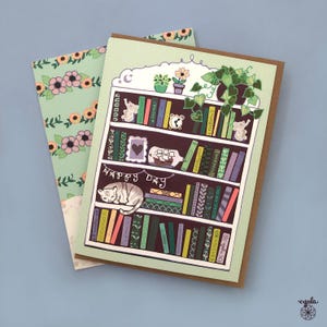 May include: Greeting card featuring a whimsical illustration of a bookshelf with books, plants, and a sleeping cat. The words "Happy Day" are written below the bookshelf. The card is paired with a floral patterned envelope.
