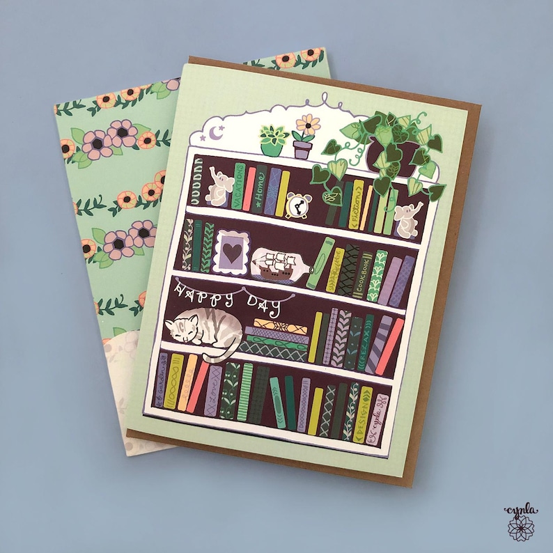 May include: Greeting card featuring a whimsical illustration of a bookshelf with books, plants, and a sleeping cat. The words "Happy Day" are written below the bookshelf. The card is paired with a floral patterned envelope.