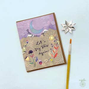 May include: A rectangular greeting card featuring a crescent moon, mountains, and floral illustrations with the text Happy New Year and Let's Try This Again. The card is accompanied by a silver star-shaped ornament and a yellow-handled paintbrush.