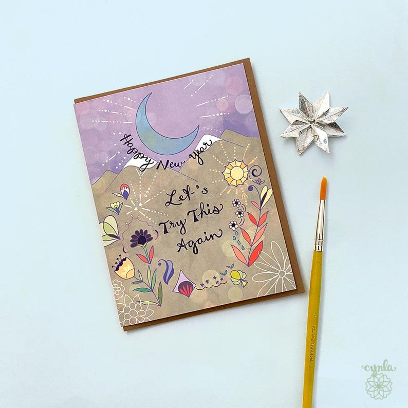 May include: A rectangular greeting card featuring a crescent moon, mountains, and floral illustrations with the text Happy New Year and Let's Try This Again. The card is accompanied by a silver star-shaped ornament and a yellow-handled paintbrush.