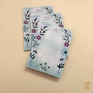 May include: Three rectangular paper notepads with a light blue gradient background. Each notepad features a floral border with purple, teal, and dark blue flowers, stars, and a crescent moon. The word notes is printed in cursive at the top center.