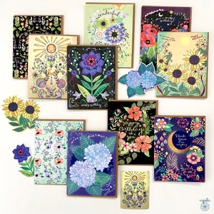May include: A collection of decorative greeting cards with floral and celestial designs. The cards feature illustrations of sunflowers, hydrangeas, and other flowers, with text such as "Happy Birthday" and "Thank you". The cards have brown borders.