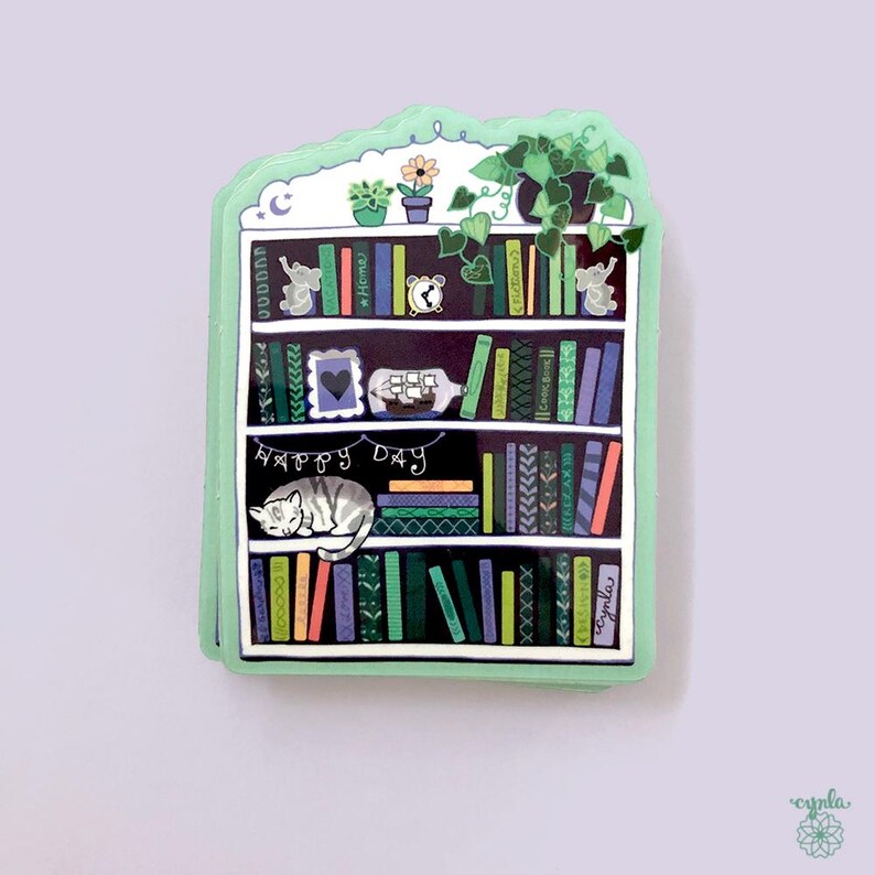 May include: A whimsical sticker in the shape of a bookshelf, featuring a sleeping cat, books, plants, and the words "Happy Day." The design includes a variety of colors and decorative elements, creating a cozy, artistic feel.