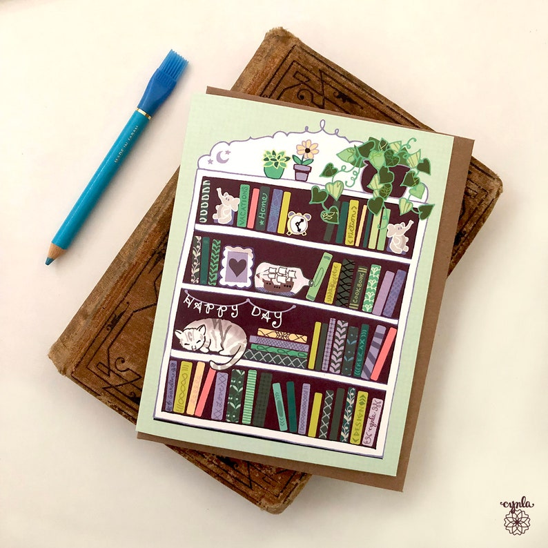 May include: A greeting card featuring a whimsical bookshelf design with a sleeping cat, books, and the text "HAPPY DAY." The card is on a brown background with a blue pencil. The card has a light green background and a brown border.