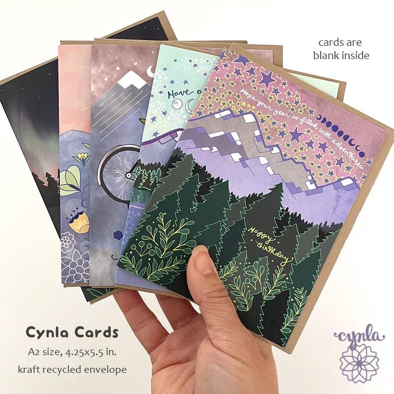 May include: A set of five greeting cards with kraft envelopes, each featuring colorful illustrations of mountains, forests, stars, and moon phases. The front card displays the text 'Hope your year is filled with adventures' and 'Happy Birthday'. Cards are 4.25x5.5 in.