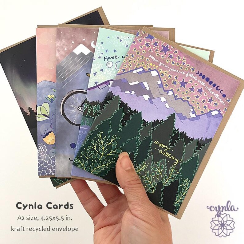 May include: A hand holds a stack of illustrated greeting cards. The cards feature nature-themed designs with mountains, trees, and a bicycle. Each card is A2 size, measuring 4.25 x 5.5 inches. The envelopes are made of kraft recycled paper. The text on the cards includes "Happy Birthday".