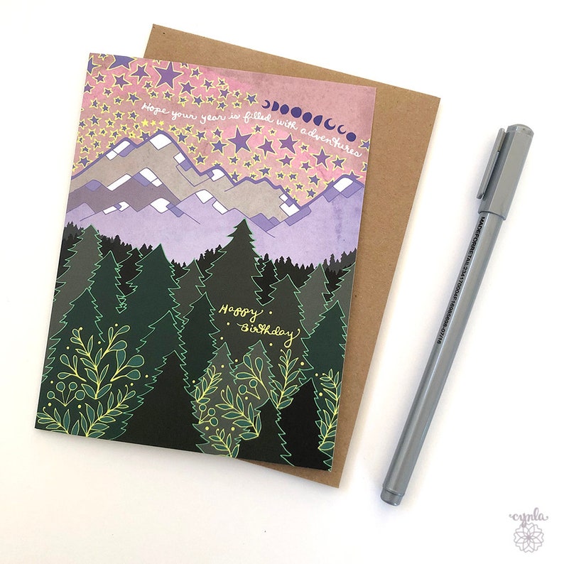May include: A greeting card with a watercolor illustration of a mountain range with a forest in the foreground. The sky is pink and purple with yellow stars and a crescent moon. The text on the card reads "Hope your year is filled with adventures" and "Happy Birthday".