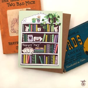May include: A greeting card featuring a bookshelf design with a sleeping cat, books, and potted plants. The card has the words "Happy Day" and is surrounded by vintage books.