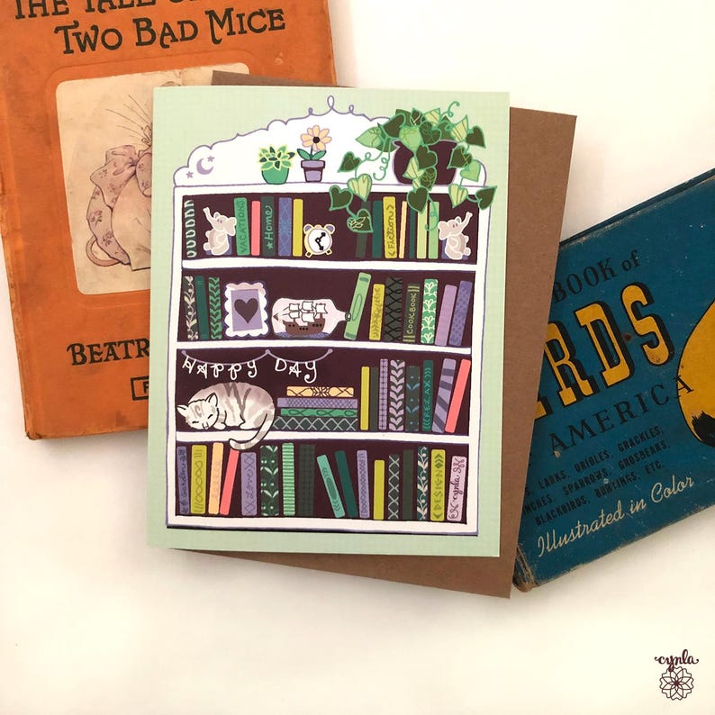 May include: A greeting card featuring a bookshelf design with a sleeping cat, books, and potted plants. The card has the words "Happy Day" and is surrounded by vintage books.