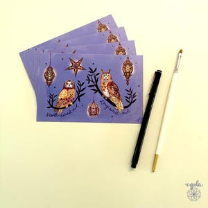 May include: A stack of rectangular postcards featuring a purple background with two owls perched on branches, a star, and hanging lanterns. The text reads Short-eared owl and Long-eared owl. A black pen and a white paintbrush with a gold tip are included.