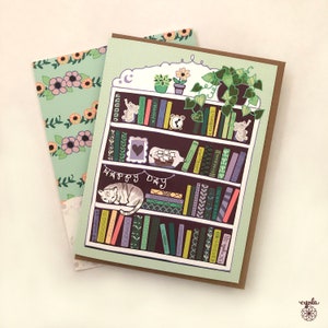 May include: A greeting card featuring a whimsical illustration of a bookshelf with books, plants, and a sleeping cat. The words "HAPPY DAY" are written across the bottom. A floral patterned envelope is behind the card.