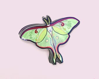 May include: A sticker featuring a detailed illustration of a Luna moth. The moth has green wings with red and purple accents, and is decorated with botanical and geometric patterns. The sticker is on a pink background.