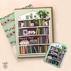 May include: A greeting card featuring a bookshelf design with a sleeping cat, books, and the words "Happy Day." The card is light green with a brown border. A smaller sticker with the same design is also present, along with a floral patterned card.
