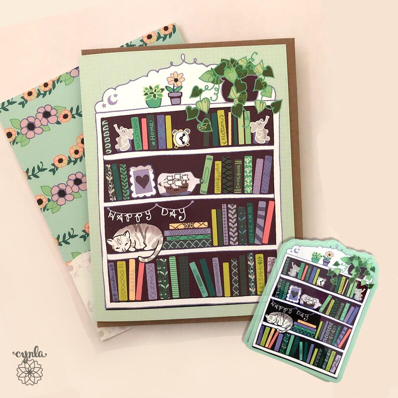 May include: A greeting card featuring a bookshelf design with a sleeping cat, books, and the words "Happy Day." The card is light green with a brown border. A smaller sticker with the same design is also present, along with a floral patterned card.