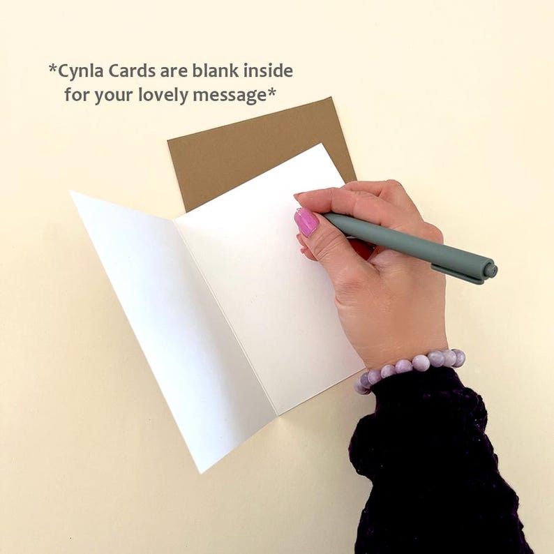 May include: An open white greeting card with a brown envelope behind it. A hand with pink nail polish holds a gray pen, writing on the blank inside of the card. The text reads, "*Cynla Cards are blank inside for your lovely message*"