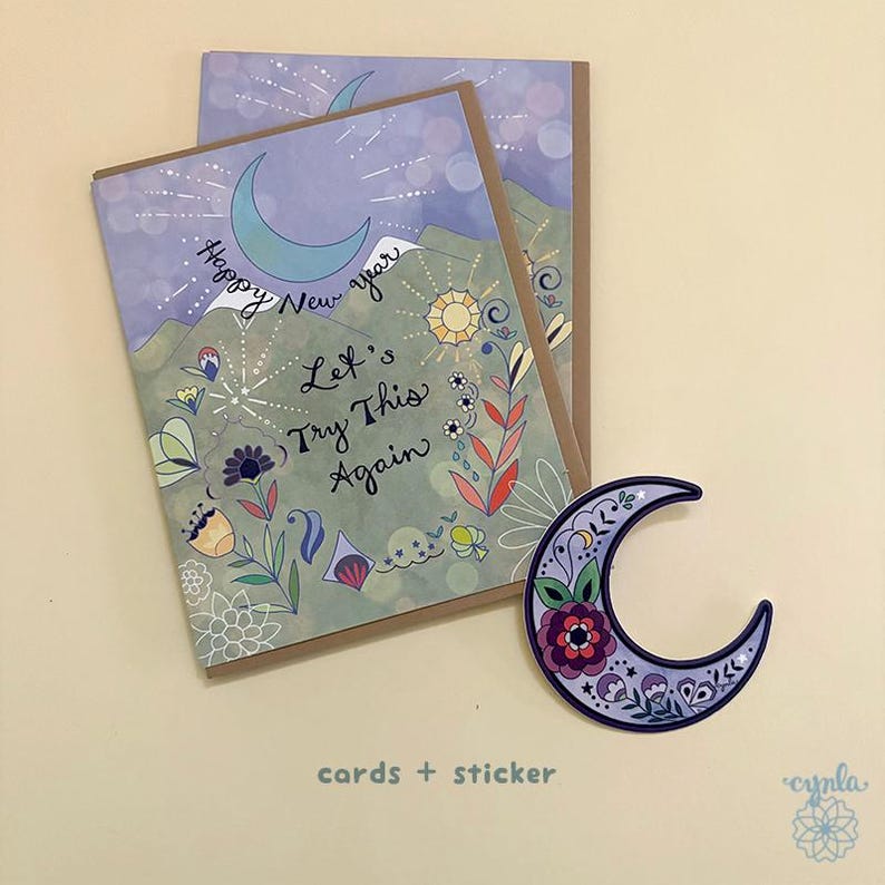 May include: Two rectangular greeting cards with a light purple and green background featuring a crescent moon, sun, and floral illustrations. The text reads Happy New Year Let's Try This Again. A crescent moon sticker with floral patterns is included.