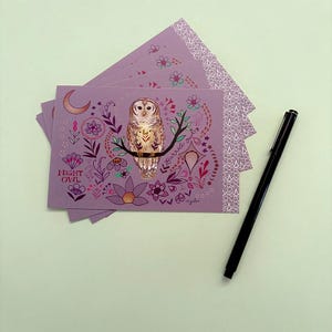 May include: A stack of rectangular purple postcards featuring a central illustration of a barred owl perched on a branch, surrounded by floral motifs, a crescent moon, and the text "NIGHT OWL" and "barred owl". A black pen lies next to the cards.