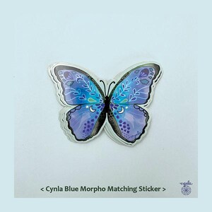 May include: A butterfly-shaped sticker featuring shades of blue and purple with white leaf patterns, small stars, and crescent moon details. The text below reads < Cynla Blue Morpho Matching Sticker > with a small circular logo on the bottom right.