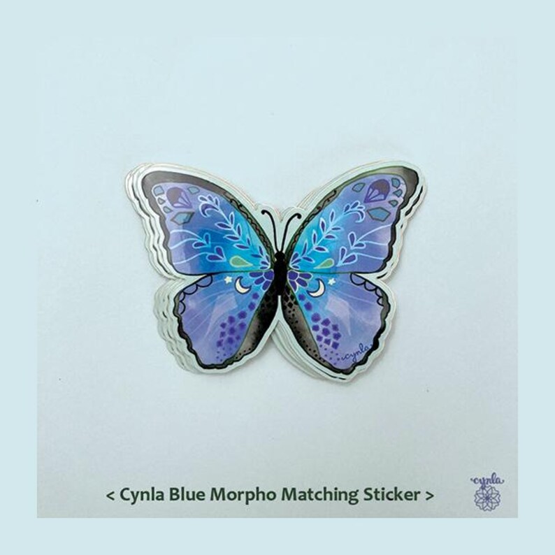 May include: A butterfly-shaped sticker featuring shades of blue and purple with white leaf patterns, small stars, and crescent moon details. The text below reads < Cynla Blue Morpho Matching Sticker > with a small circular logo on the bottom right.