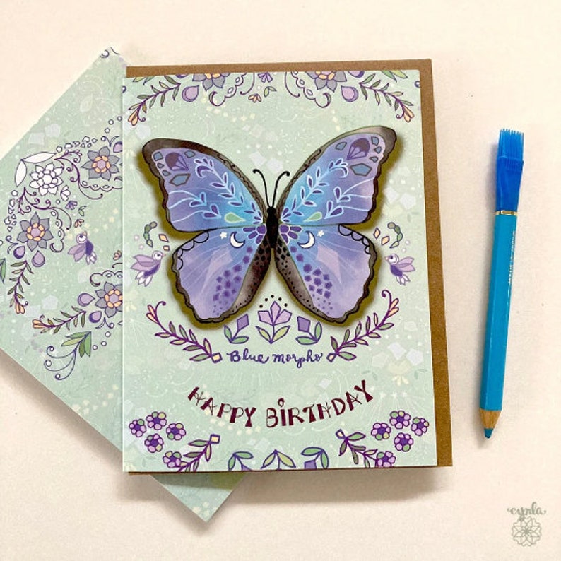 May include: A rectangular greeting card featuring a large, central illustration of a blue and purple butterfly with white crescent moon accents. The card displays the text "Blue morpho" and "HAPPY BIRTHDAY" in dark purple, surrounded by floral motifs.