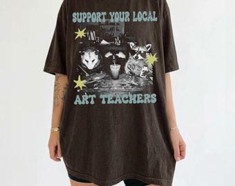 May include: A dark brown short-sleeve t-shirt featuring a graphic print of an opossum and a raccoon wearing sunglasses at a desk. The text reads SUPPORT YOUR LOCAL ART TEACHERS in light blue, accented by two yellow star shapes on the sides.