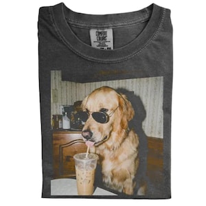 May include: A charcoal grey crew neck t-shirt featuring a graphic print of a golden retriever wearing black aviator sunglasses. The dog is positioned next to a clear plastic cup filled with an iced beverage and a straw, set against a room interior.