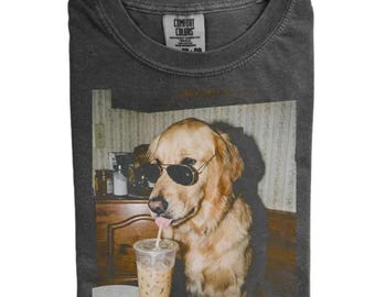 Puede incluir: Una camiseta de cuello redondo gris carbón con un estampado gráfico de un golden retriever que lleva gafas de sol de aviador negras. El perro está situado junto a un vaso de plástico transparente lleno de una bebida helada y una pajita.