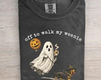 May include: A charcoal gray crew neck t-shirt featuring a graphic of a white ghost holding a pumpkin balloon and a coffee cup, walking a dachshund wearing a witch hat. The text reads off to walk my weenie in a white arched font above the graphic.