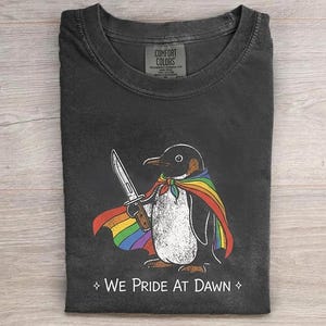 May include: A charcoal gray crew neck t-shirt featuring a graphic of a penguin wearing a rainbow-colored cape and holding a knife. Below the penguin, the text reads "WE PRIDE AT DAWN" with a small four-pointed star symbol on each side of the phrase.