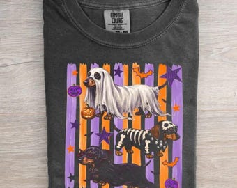May include: A charcoal gray t-shirt featuring a graphic of three dachshunds in Halloween costumes. One dog wears a ghost sheet, one has a skeleton print, and one is dressed as a witch. The background has orange and purple vertical stripes.
