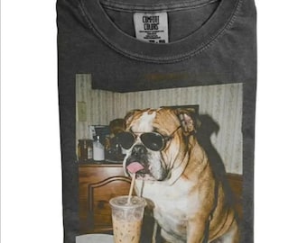 May include: A charcoal gray crew neck t-shirt featuring a graphic print of a bulldog wearing black aviator sunglasses and holding a pink lollipop in its mouth while sitting next to a clear plastic cup filled with an iced coffee beverage.