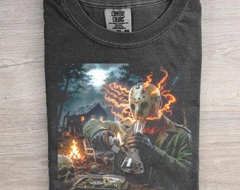 May include: A charcoal gray crew neck t-shirt featuring a graphic print of a hockey-masked figure wearing a green jacket, surrounded by orange flames, holding a glass water pipe. The background shows a cabin at night with a campfire and a skull.