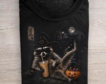 May include: A black crew neck t-shirt featuring a graphic of a raccoon wearing a witch hat and sunglasses, holding an iced coffee. Beside the raccoon is a jack-o'-lantern bucket. The text "LIVE FAST EAT TRASH" is printed on a small sign.