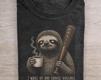 May include: A black crew neck t-shirt featuring a graphic of a sloth holding a steaming coffee mug and a barbed wire wrapped baseball bat. The text below the sloth reads, I WAKE UP AND CHOOSE VIOLENCE. The design is rendered in shades of brown and white.