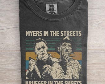 May include: A charcoal gray crew neck t-shirt featuring a graphic print of two horror movie characters. The text reads MYERS IN THE STREETS KRUEGER IN THE SHEETS. The design includes a retro-style horizontal striped background in muted colors.