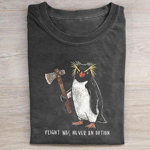May include: A charcoal gray crew neck t-shirt featuring a graphic of a penguin with yellow crest feathers holding a brown axe. Below the penguin, the text reads: FLIGHT WAS NEVER AN OPTION. The shirt has short sleeves and a standard collar design.