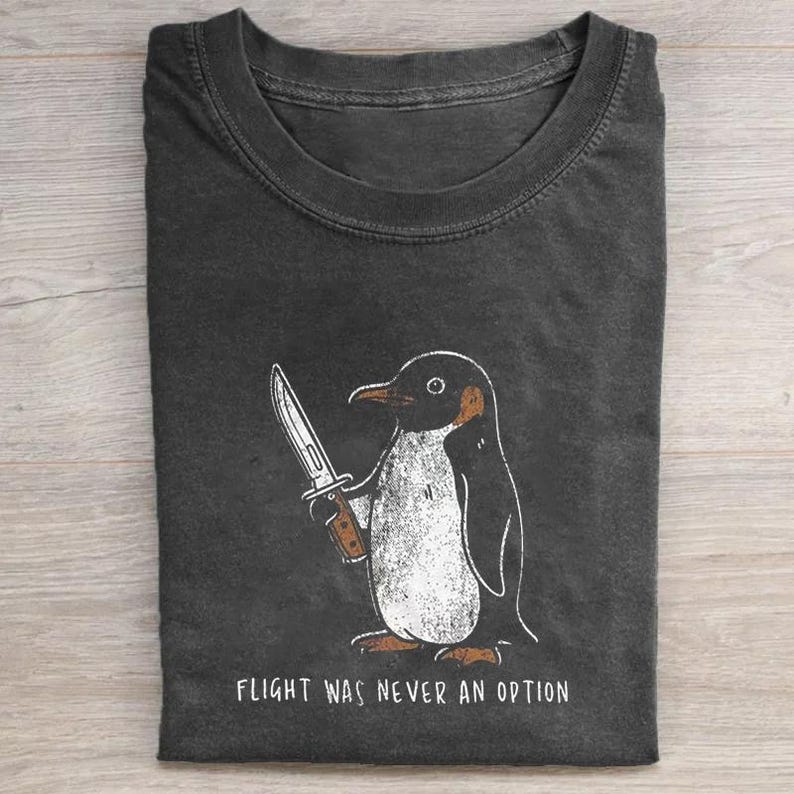 Puede incluir: Camiseta gris oscuro con un dibujo de un ping&uuml;ino sosteniendo un cuchillo. El ping&uuml;ino es negro, blanco y marr&oacute;n. El texto "FLIGHT WAS NEVER AN OPTION" est&aacute; impreso debajo.