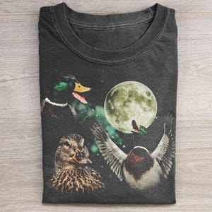 May include: Dark gray t-shirt featuring a whimsical design of ducks, a full moon, and butterflies. The ducks are depicted in various poses, with one appearing to be quacking. The moon is a soft green color, and the butterflies add a touch of fantasy.