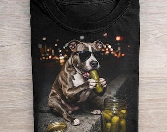 May include: A black t-shirt featuring a graphic of a brown and white dog wearing black sunglasses. The dog is holding a green pickle in its paws, with a glass jar filled with pickles sitting on the ground next to it. The background shows city lights.