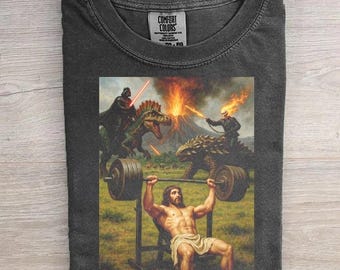 May include: A charcoal gray crew neck t-shirt featuring a graphic print of a person lifting a barbell on a bench press. In the background, a figure rides a dinosaur toward a volcanic eruption while another figure holds a flaming chain.