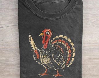 May include: A charcoal gray crew neck t-shirt featuring a graphic of a turkey holding a knife in its wing. The turkey has red and orange plumage details. Below the graphic, the text reads: THANKSGIVING WAS NEVER AN OPTION.