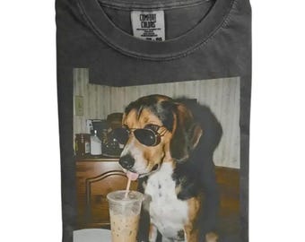May include: A charcoal gray t-shirt featuring a graphic print of a beagle wearing black sunglasses and drinking from a clear plastic cup with a straw. The shirt has a Comfort Colors brand label on the inside of the collar and a crew neckline.