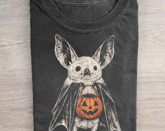 Puede incluir: Una camiseta gris carbón con una ilustración gráfica de un murciélago de color claro con orejas grandes y colmillos. El murciélago aparece con las alas parcialmente extendidas, sosteniendo un cubo de calabaza naranja brillante.