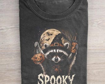 May include: A charcoal gray t-shirt featuring a graphic of a raccoon wearing a witch hat and heart-shaped sunglasses. The raccoon holds a jack-o'-lantern and a coffee cup against a full moon background. The word SPOOKY is printed in white below.