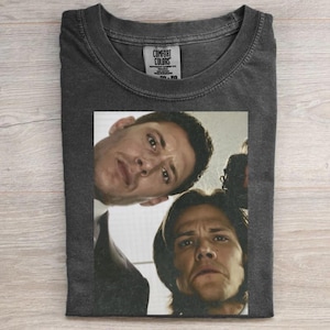 May include: A charcoal gray crew neck t-shirt featuring a graphic print of two men looking downward from a high angle. The shirt has a Comfort Colors brand label inside the collar. The fabric has a washed, vintage appearance with a soft texture.