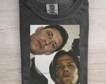 May include: A charcoal gray crew neck t-shirt featuring a graphic print of two men looking downward from a high angle. The shirt has a Comfort Colors brand label inside the collar. The fabric has a washed, vintage appearance with a soft texture.