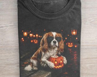 May include: A charcoal gray t-shirt featuring a graphic of a brown and white Cavalier King Charles Spaniel wearing black sunglasses. The dog is holding a small, glowing jack-o'-lantern with a whipped cream topping against a dark, bokeh background.